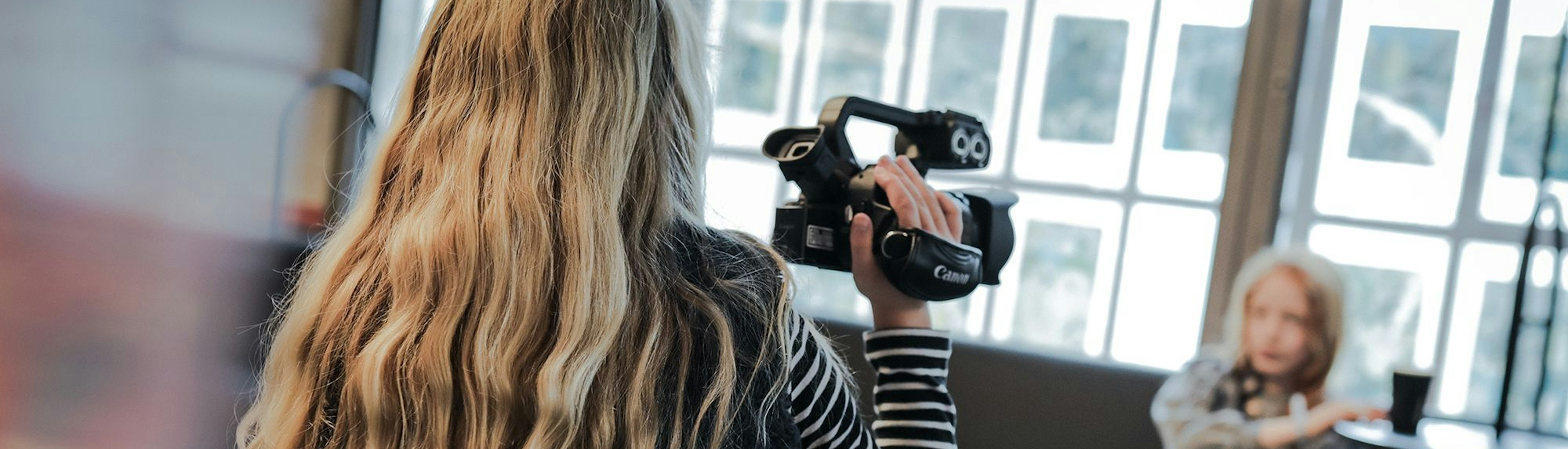 Eine Person mit langen, welligen blonden Haaren filmt ein kleines Kind, das auf einer gepolsterten Bank vor einem großen Fenster sitzt. Das Kind gestikuliert beim Sprechen mit einer Hand.