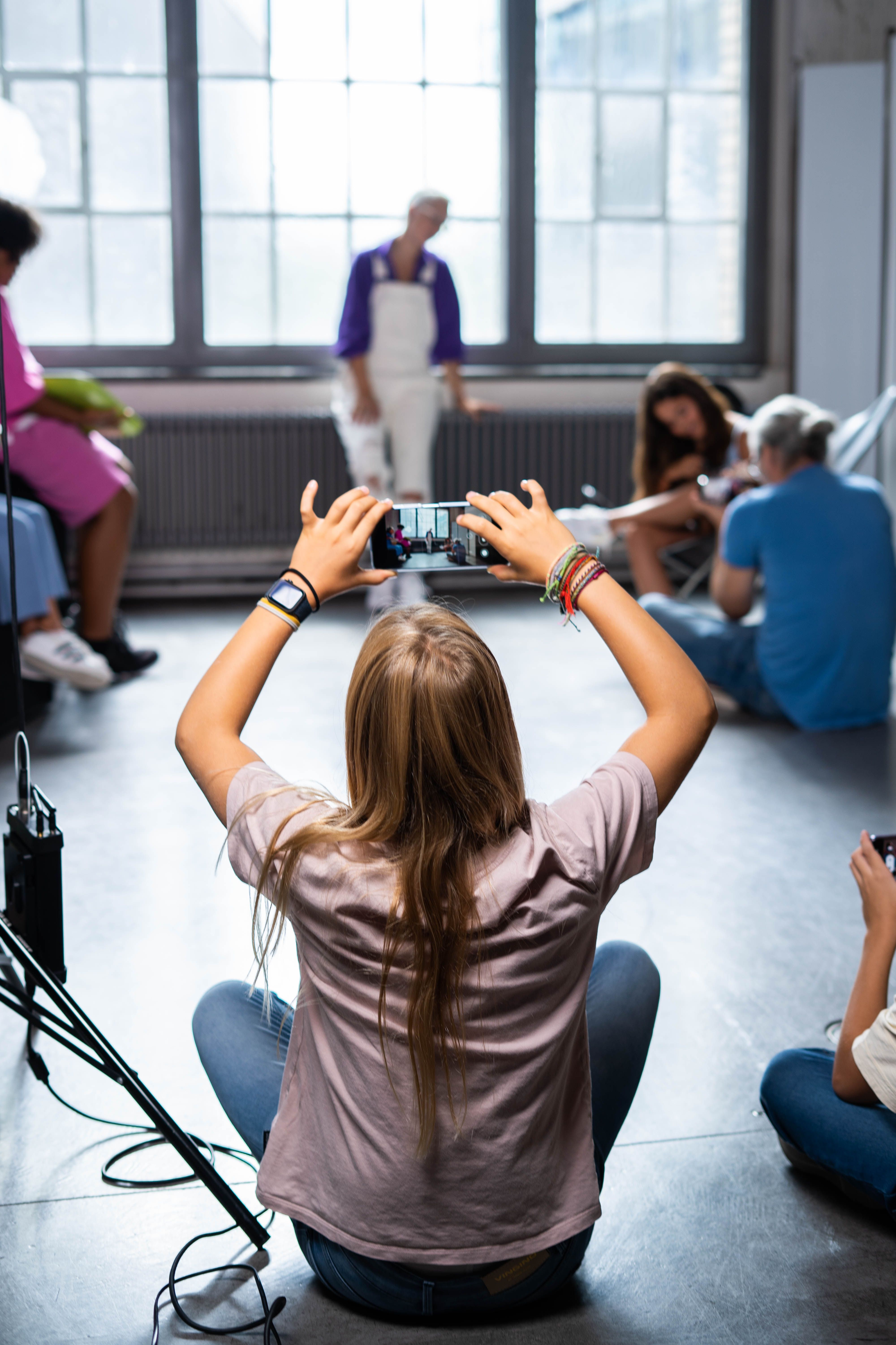 Eine Person sitzt auf dem Boden und filmt oder fotografiert eine Gruppe vor großen Fenstern. Dabei wird eine stehende Person fokussiert, während andere in der Nähe sitzen und sich unterhalten. Die Szenerie scheint ein kreativer oder lockerer Workshop zu sein.