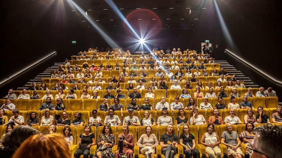 Ein volles Publikum sitzt in gelben Theatersitzen mit Blick auf die Bühne, die von einem hellen Scheinwerfer aus dem hinteren Teil des Saals beleuchtet wird. Der Raum ist dunkel, und das Publikum wirkt aufmerksam.