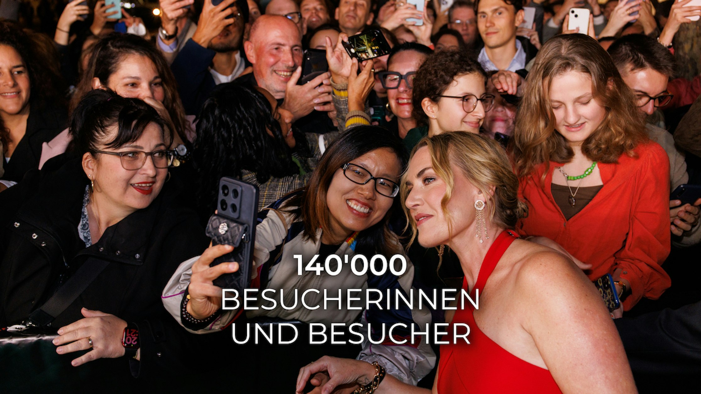 Eine Frau im roten Kleid macht ein Selfie mit lächelnden Fans in einer Menschenmenge. Viele Menschen halten ihre Handys hoch, um den Moment festzuhalten. Weißer Text auf dem Bild lautet: 140000 BESUCHERINNEN UND BESUCHER.