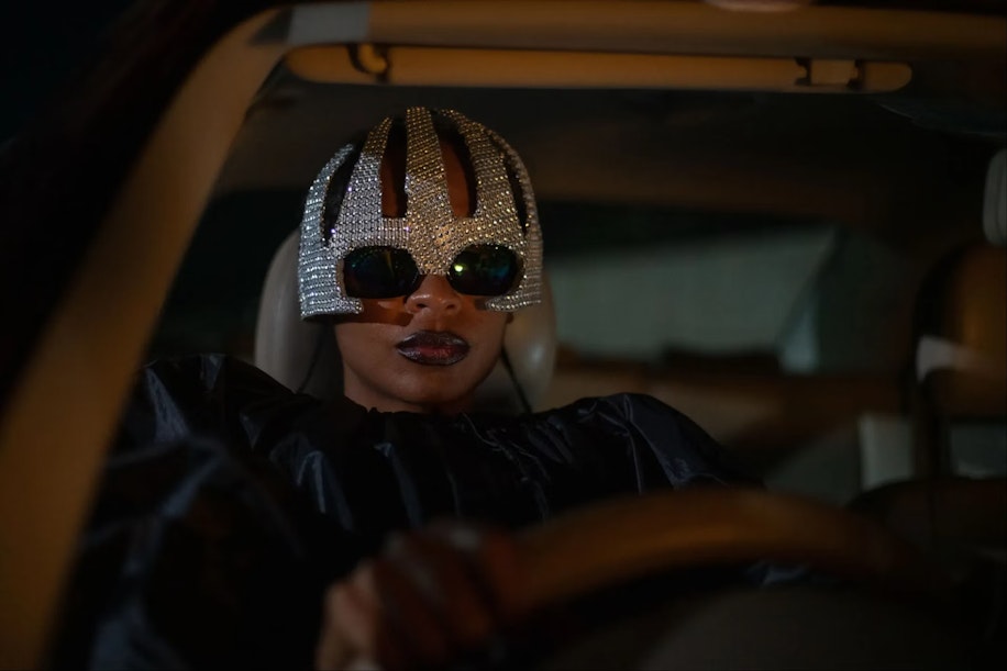 Eine Person mit einer glitzernden, mit Juwelen besetzten Maske und einer dunklen Sonnenbrille sitzt nachts auf dem Fahrersitz eines Autos, dramatisch beleuchtet, mit ernstem Gesichtsausdruck und den Händen am Lenkrad.