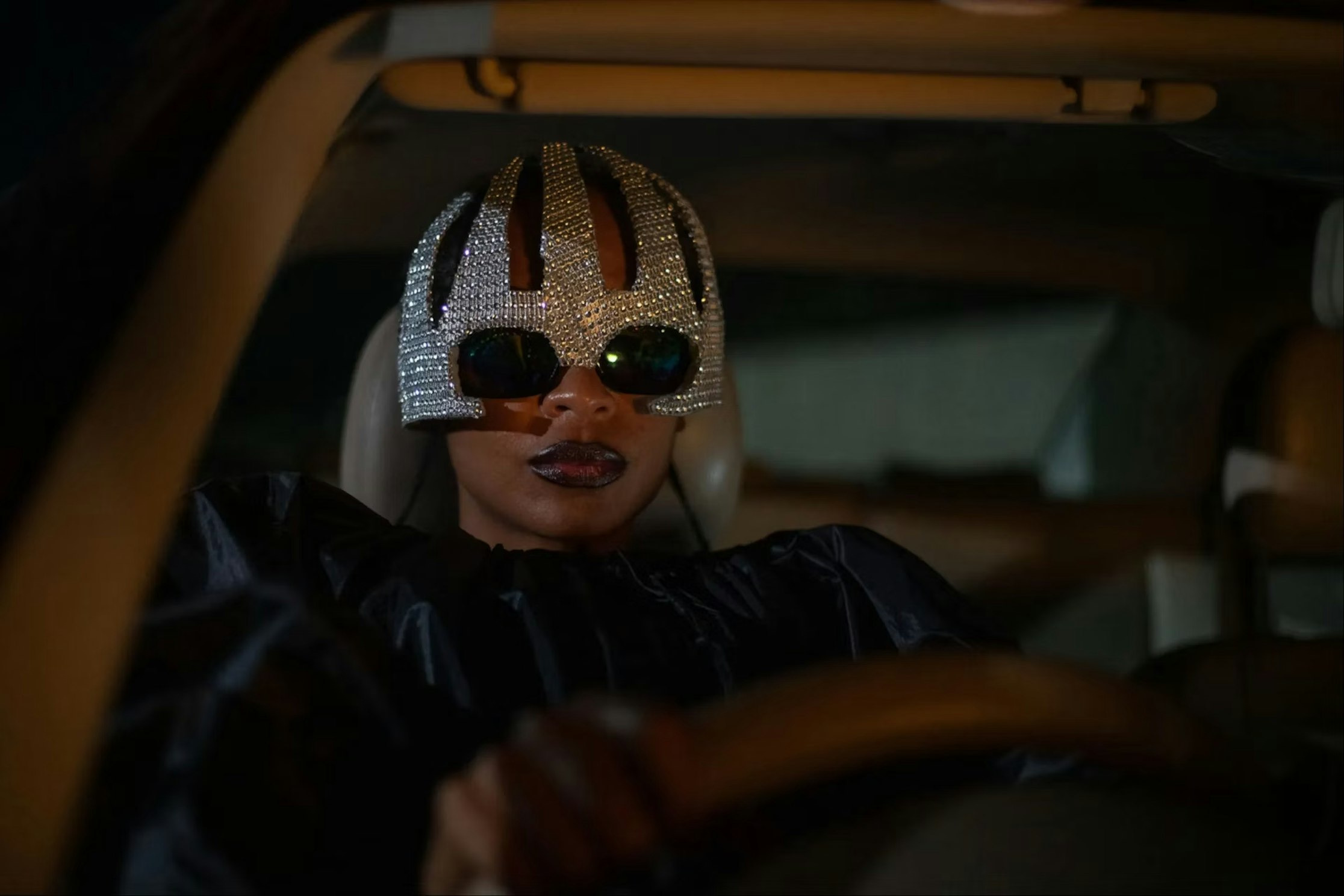 Eine Person mit einer glitzernden, mit Juwelen besetzten Maske und einer dunklen Sonnenbrille sitzt nachts auf dem Fahrersitz eines Autos, dramatisch beleuchtet, mit ernstem Gesichtsausdruck und den Händen am Lenkrad.