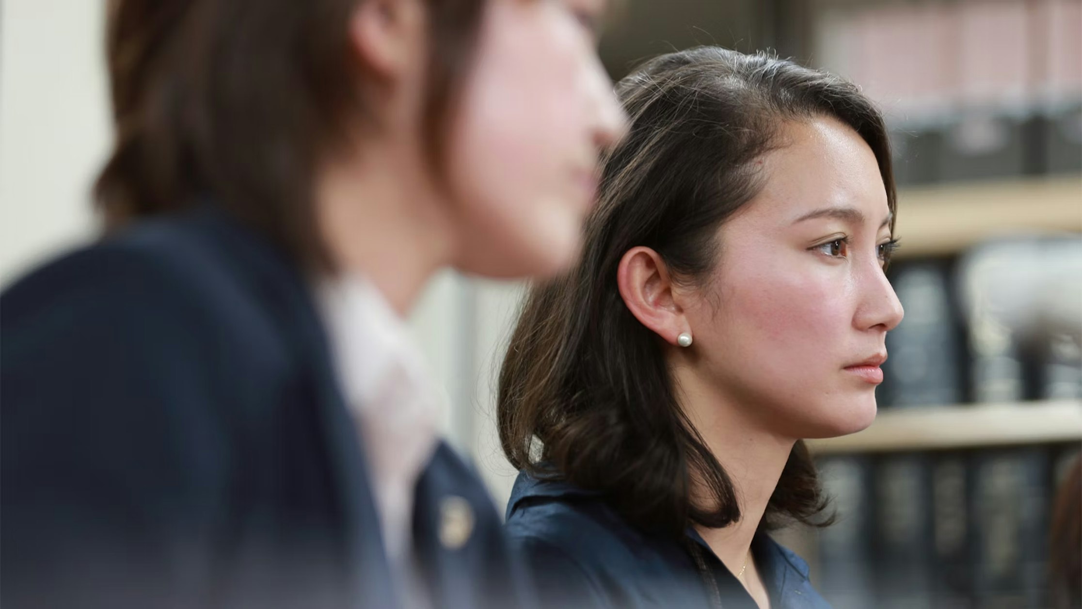 Zwei Frauen in Businesskleidung sitzen im Innenbereich; die eine ist scharf zu sehen und blickt nachdenklich geradeaus, die andere ist verschwommen im Vordergrund zu sehen. Im Hintergrund sind Regale mit Büchern zu sehen.