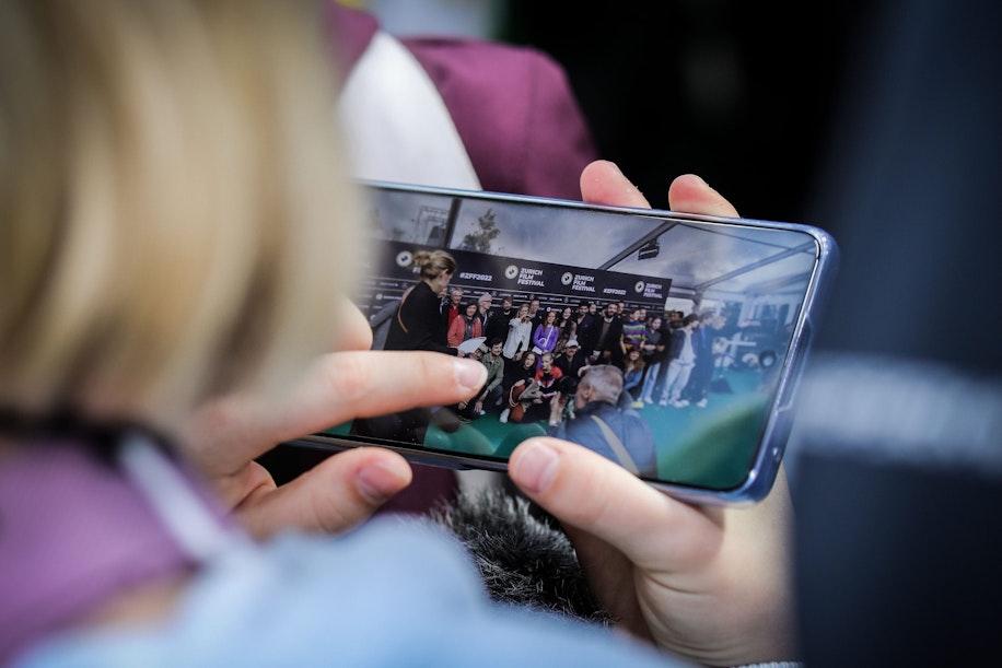Eine Person hält ein Smartphone und betrachtet ein Gruppenfoto auf dem Bildschirm. Das Bild zeigt mehrere Personen, die vor einem Hintergrund mit Logos posieren. Die Hand der Person berührt teilweise den Bildschirm.