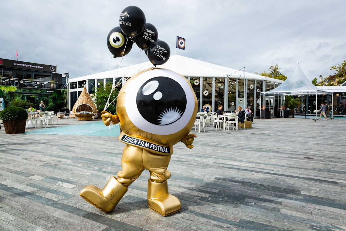 A gold mascot with a large eye for a head holds black Zurich Film Festival balloons in an outdoor plaza, with a modern tent structure and seating area in the background.