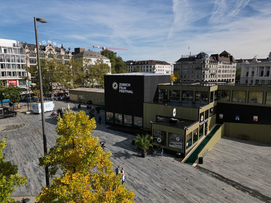 Ein Blick auf das Gelände des Zurich Film Festival mit schwarzen und grünen Gebäuden, Sitzgelegenheiten im Freien und Bäumen in der Nähe, gelegen auf einem städtischen Platz mit historischen Gebäuden im Hintergrund.