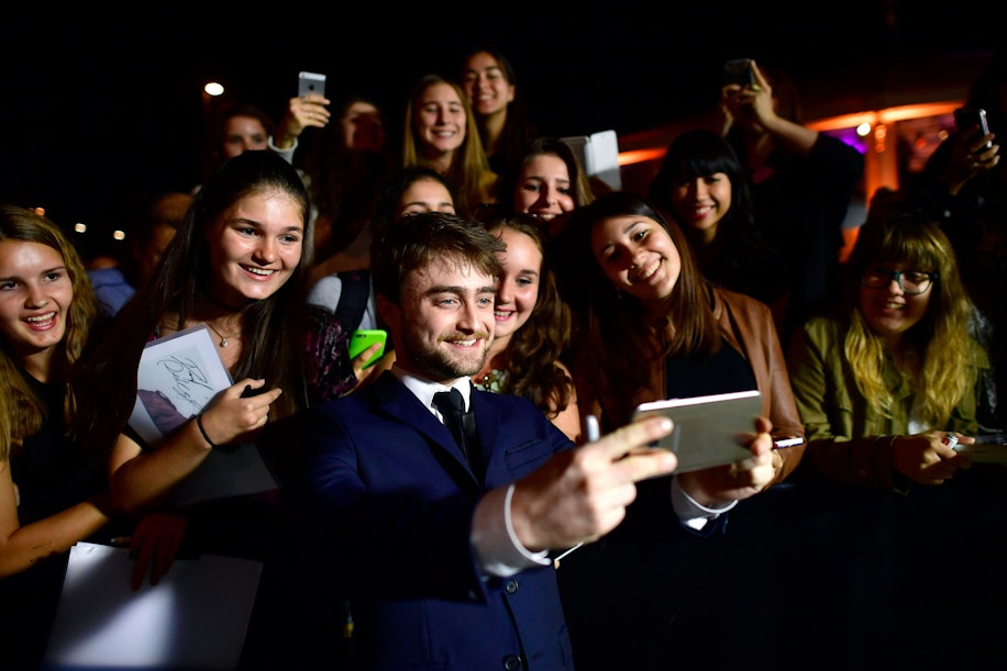 Ein Mann im Anzug macht bei einer gut besuchten Veranstaltung, wahrscheinlich nachts, ein Selfie mit einer Gruppe lächelnder Fans, die Telefone und Papiere in den Händen halten.