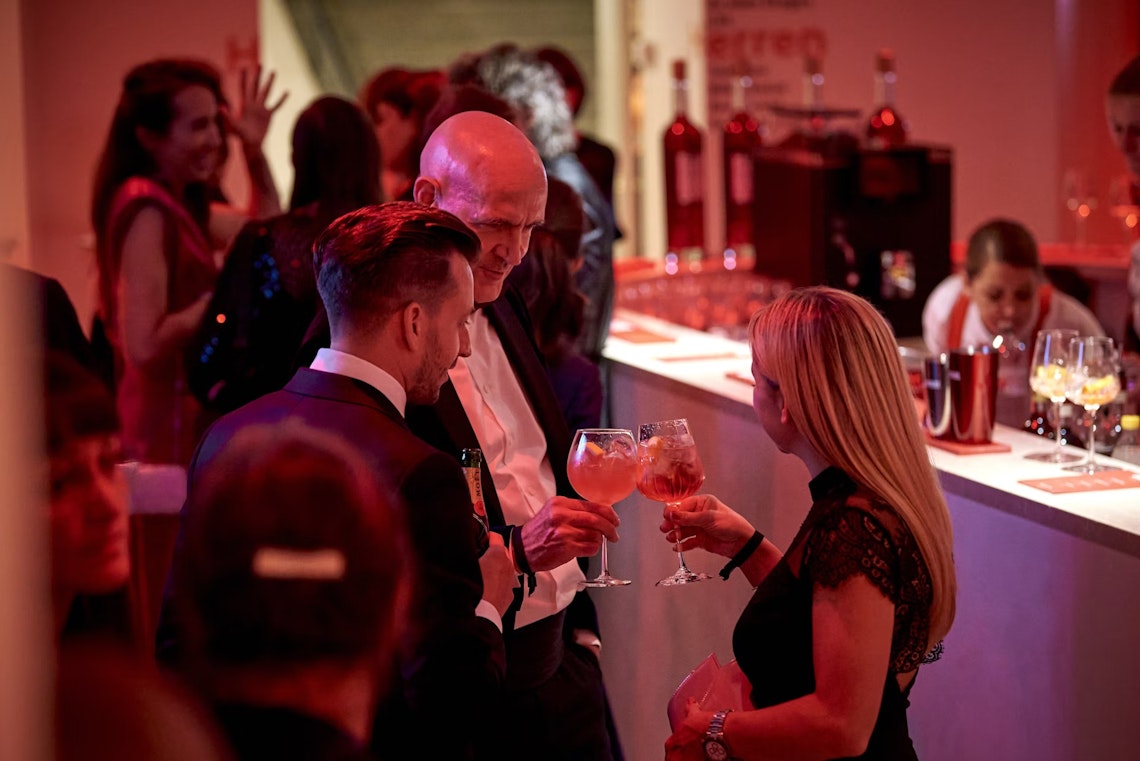 People in formal attire converse and hold drinks at a modern, dimly lit bar with red lighting. Bottles and glasses are visible on the counter, and others mingle in the background.