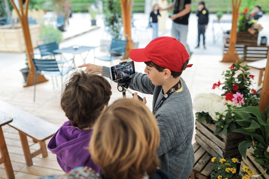 Ein Junge mit einer roten Mütze zeigt zwei anderen Kindern in einer hölzernen Sitzecke im Freien, umgeben von Blumen und Pflanzen, etwas auf einem auf einem Stativ montierten Smartphone.