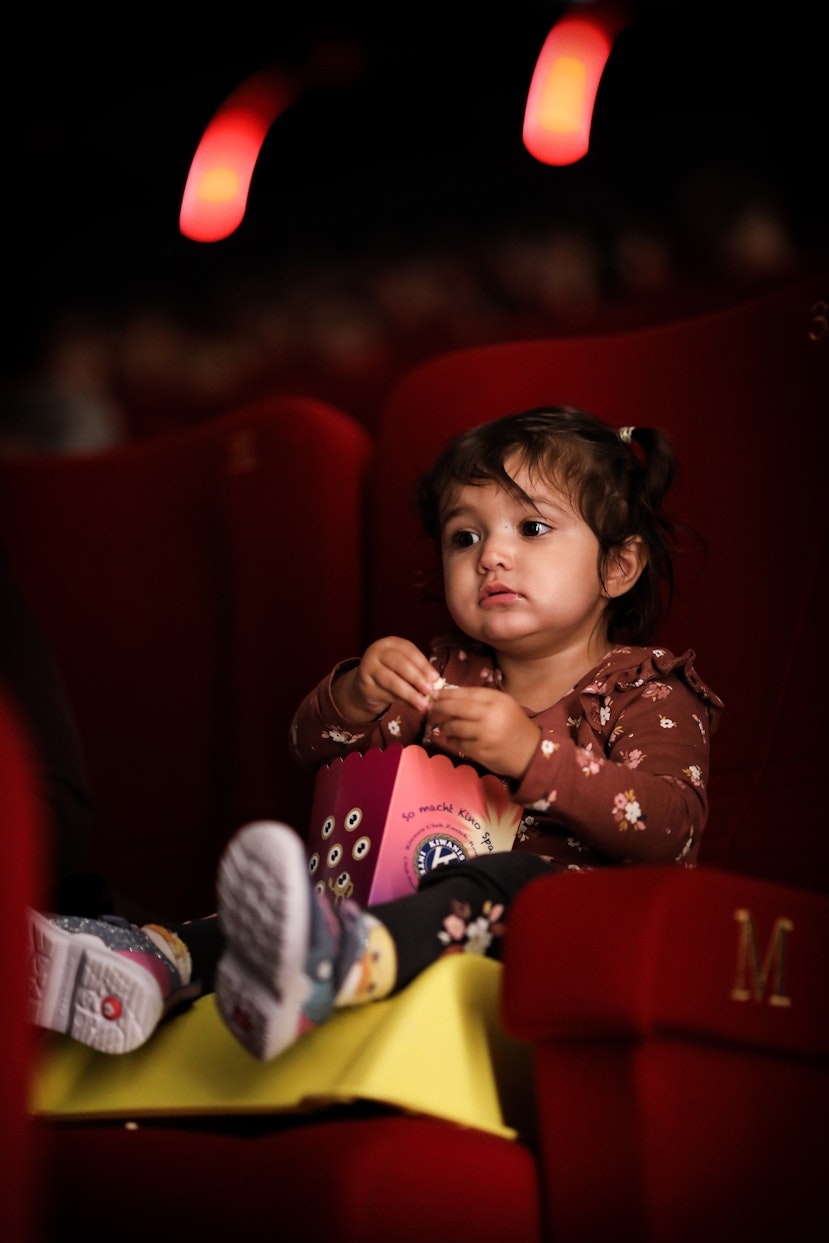 Ein kleines Kind sitzt allein in einem roten Kinosessel und isst Popcorn mit nachdenklichem Gesichtsausdruck. Der Hintergrund ist dunkel und sanft rot beleuchtet, was darauf schließen lässt, dass es einen Film sieht.
