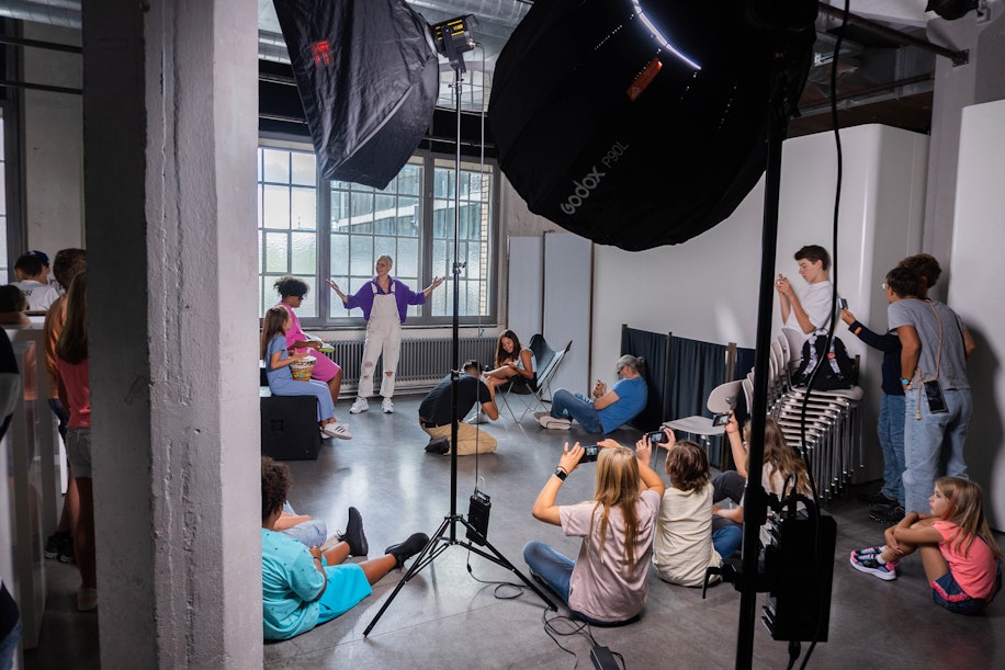 Eine Gruppe von Menschen in einem Fotostudio beobachtet eine Person, die mit ausgestreckten Armen am Fenster steht und posiert. Mehrere Personen sitzen oder knien und fotografieren, während Studiolichter und -ausrüstung die Szene umgeben.