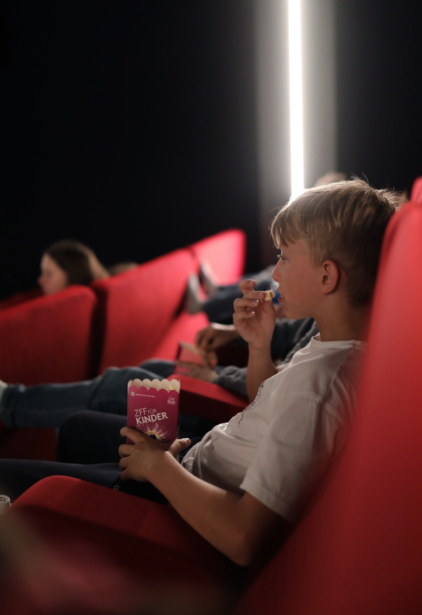 Ein Junge sitzt in einem roten Kinosessel und isst Snacks, während er einen Film sieht. Andere Kinder sitzen in der Nähe, und der Raum ist schwach beleuchtet, im Hintergrund ist ein Lichtstreifen zu sehen.