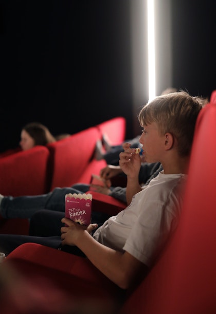 Ein Junge sitzt in einem roten Kinosessel und isst Snacks, während er einen Film sieht. Andere Kinder sitzen in der Nähe, und der Raum ist schwach beleuchtet, im Hintergrund ist ein Lichtstreifen zu sehen.