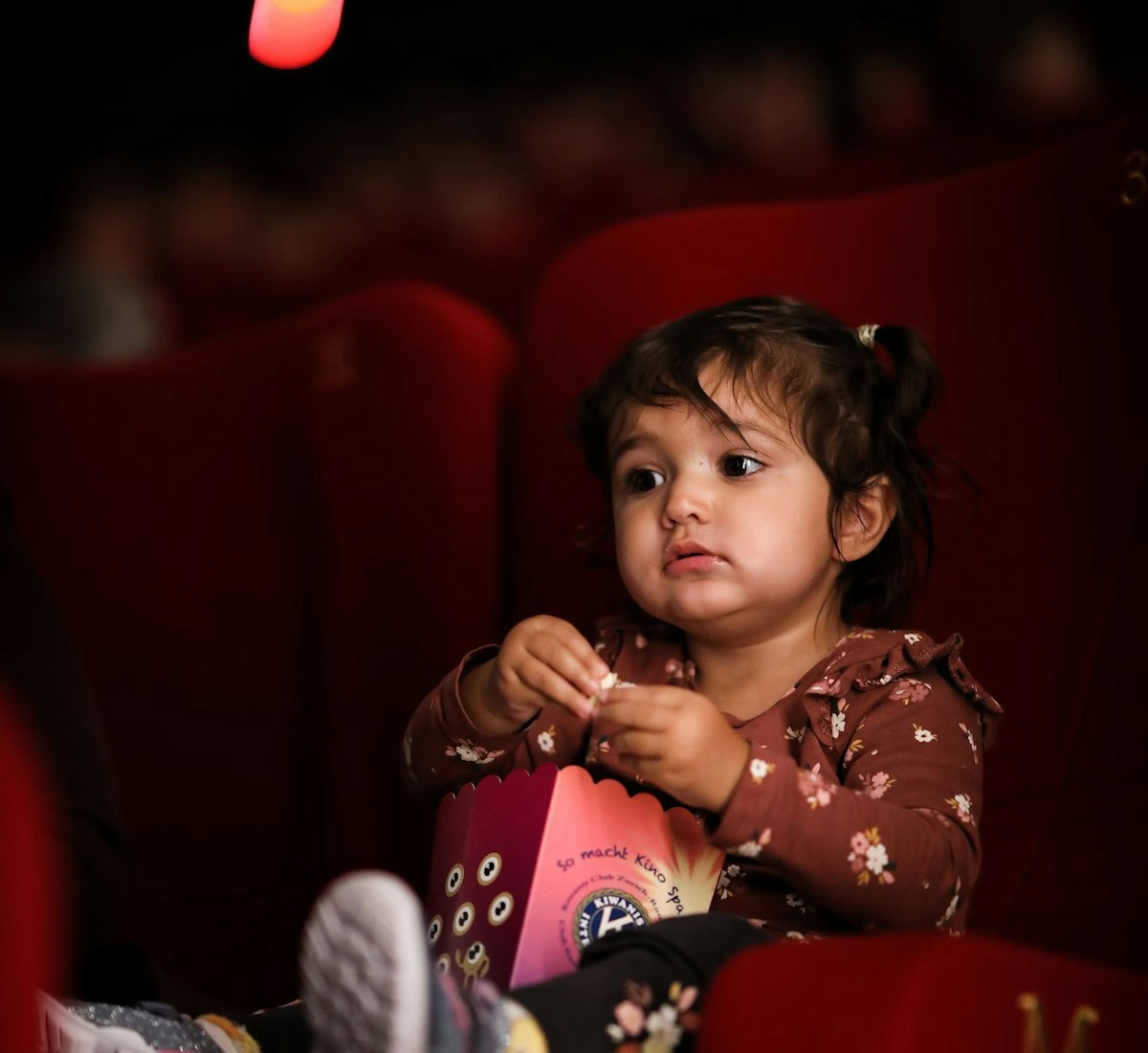 zff_studio_premiere Ein kleines Kind sitzt in einem roten Kinositz, hält eine Popcorndose in der Hand und schaut aufmerksam zu. Es trägt ein braunes Oberteil mit Blumenmuster und bunte Socken. Sanftes Licht hebt sein Gesicht vor einem dunklen Hintergrund hervor.