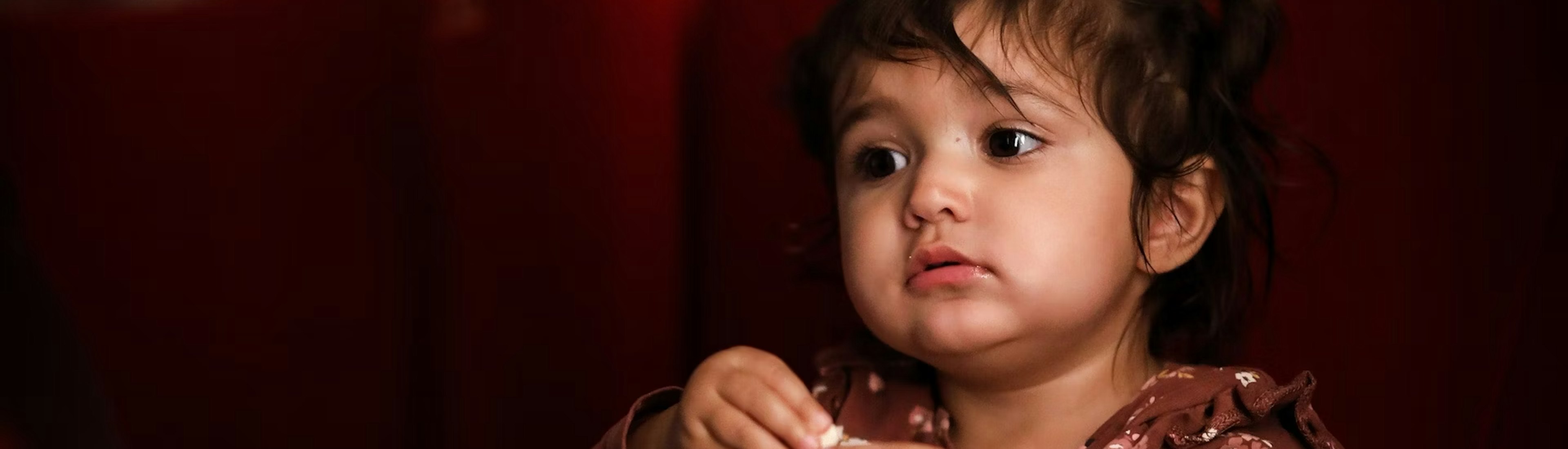 zff_studio_premiere Ein kleines Kind sitzt in einem roten Kinositz, hält eine Popcorndose in der Hand und schaut aufmerksam zu. Es trägt ein braunes Oberteil mit Blumenmuster und bunte Socken. Sanftes Licht hebt sein Gesicht vor einem dunklen Hintergrund hervor.