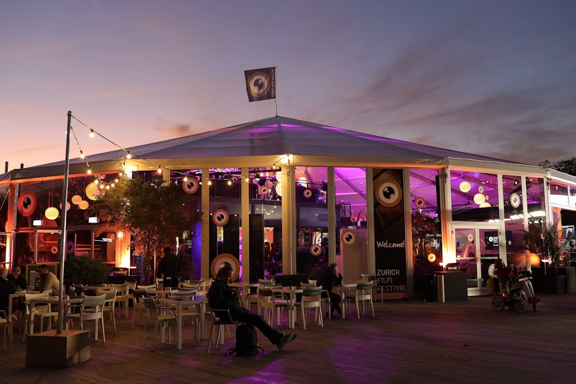 Outdoor seating area at dusk with a large, well-lit tent decorated with round eye-like signs, purple lighting, and a flag on top, likely hosting the Zurich Film Festival. People are seated and relaxing.
