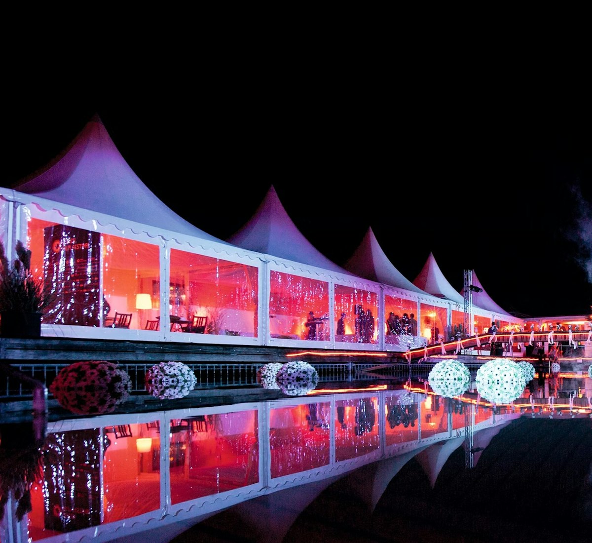 zurich_film_festival_hospitality_frauenbadi Große weiße Zelte mit spitzen Dächern werden nachts hell mit rosa und violetten Lichtern beleuchtet, die sich in einem ruhigen Teich spiegeln. Dekorative Kugeln schwimmen auf dem Wasser, und Nebel steigt am Rand auf und schafft eine festliche Atmosphäre.