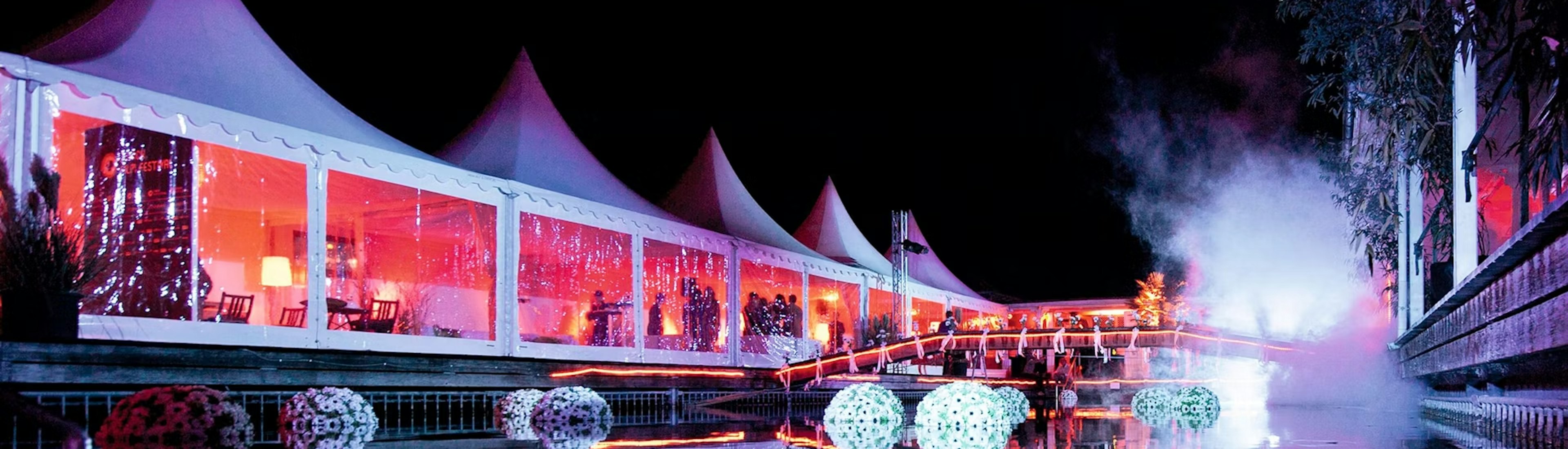 zurich_film_festival_hospitality_frauenbadi Große weiße Zelte mit spitzen Dächern werden nachts hell mit rosa und violetten Lichtern beleuchtet, die sich in einem ruhigen Teich spiegeln. Dekorative Kugeln schwimmen auf dem Wasser, und Nebel steigt am Rand auf und schafft eine festliche Atmosphäre.