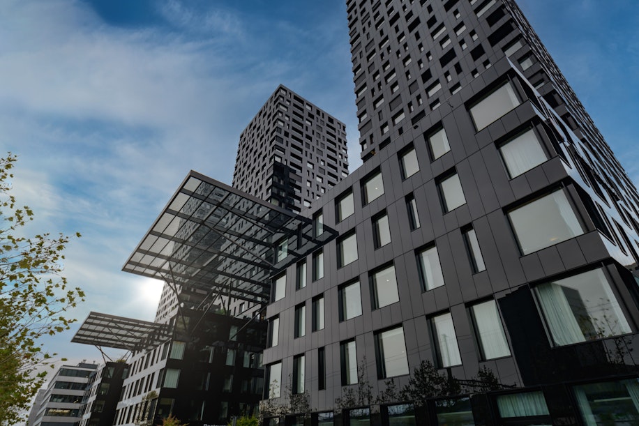 Moderne schwarze Hochhäuser mit unregelmäßigen Fenstermustern, Glasdächern und einem blauen, teilweise bewölkten Himmel im Hintergrund. Am Fuße der Gebäude ist etwas Grün zu sehen.