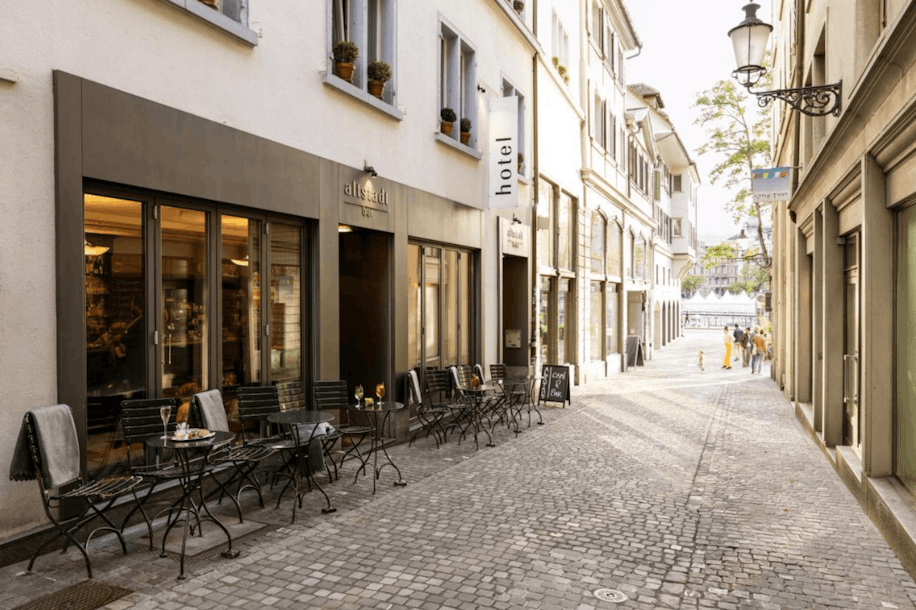 Eine schmale, gepflasterte Straße mit Tischen und Stühlen im Freien neben einem modernen Restaurant und Hotel; die Gebäude haben große Fenster und darüber leuchtet eine klassische Straßenlaterne.