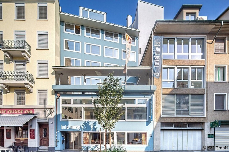 Vorderansicht des Hotels Marta in Zürich, einem modernen blau-grauen Gebäude mit großen Fenstern, eingebettet zwischen älteren Gebäuden, darunter links ein Pub. Vor dem Hoteleingang steht ein kleiner Baum.