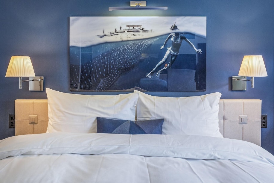 Ein ordentlich gemachtes Bett mit weißer Bettwäsche und Kissen steht vor einer blauen Wand, flankiert von zwei Wandlampen. Über dem Bett hängt ein gerahmtes Kunstwerk, das einen Schwimmer und einen Walhai unter Wasser zeigt.
