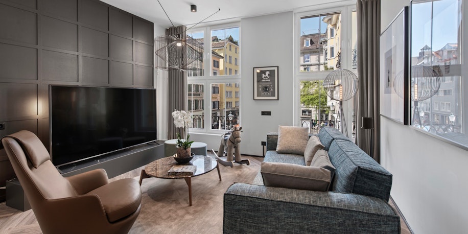 Modernes Wohnzimmer mit großem Fernseher, blauem Sofa, braunem Sessel, rundem Couchtisch aus Holz, Stehlampe und großen Fenstern, die Tageslicht hereinlassen und den Blick auf die umliegenden Gebäude freigeben. Neutrale Töne und minimalistisches Dekor.