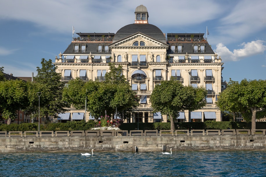 Ein prächtiges, historisches Gebäude mit kunstvoller Architektur und blau gestreiften Fenstermarkisen steht am Wasser, teilweise im Schatten grüner Bäume. Im Vordergrund schwimmen zwei Schwäne auf dem Steindamm.