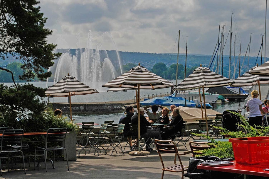 Menschen sitzen an Tischen im Freien unter gestreiften Sonnenschirmen in der Nähe eines Sees mit Segelbooten und einem großen Brunnen im Hintergrund. Im Vordergrund sind einige Bäume und ein paar leere Stühle zu sehen. Darüber ist der Himmel bewölkt.