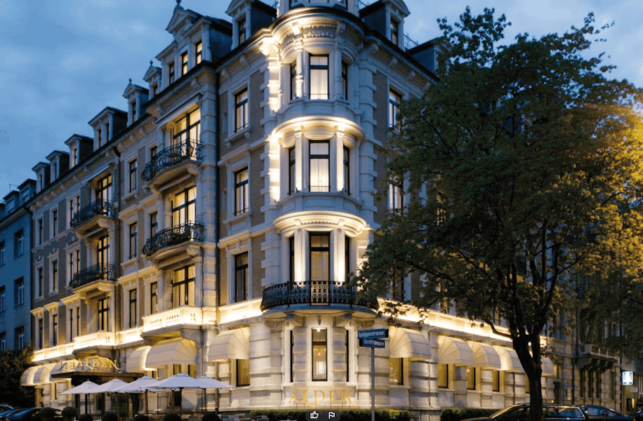 Elegantes, historisches mehrstöckiges Gebäude, dessen Fassade in der Dämmerung von warmem Licht erhellt wird; große Fenster, Balkone und ein Restaurant im Erdgeschoss mit Sitzgelegenheiten im Freien; Bäume rahmen die Ecklage an einer Stadtstraße ein.