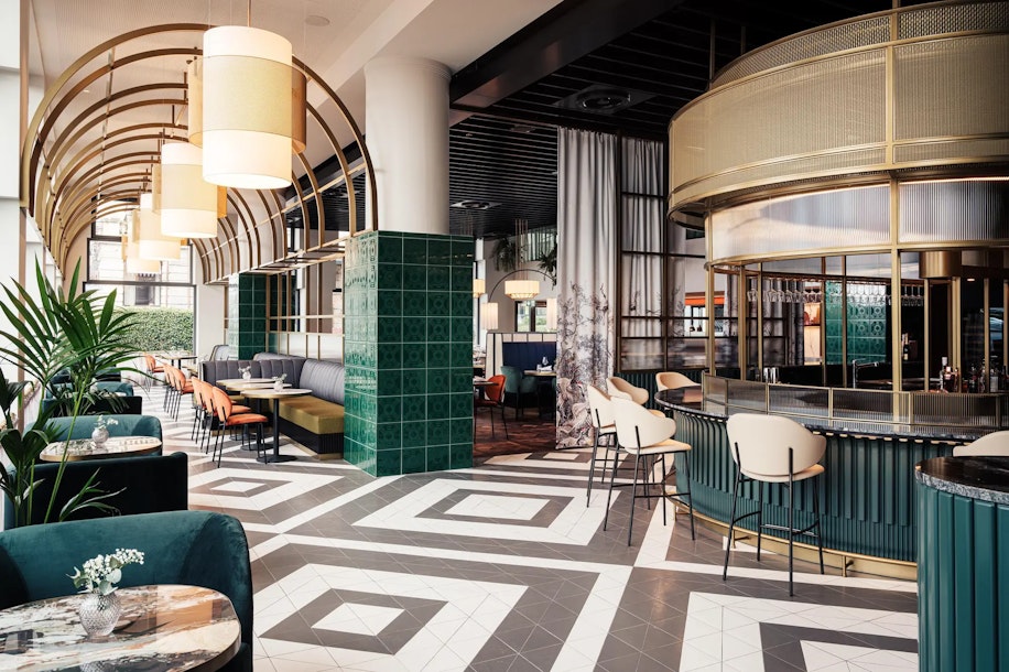 Modernes, stilvolles Restaurant-Interieur mit geometrischen Fliesenböden, grünen und goldenen Akzenten, geschwungenen Bogendetails, gepolsterten Sitzgelegenheiten, runden Tischen, Barbereich und großen Hängelampen, die eine einladende und elegante Atmosphäre schaffen.