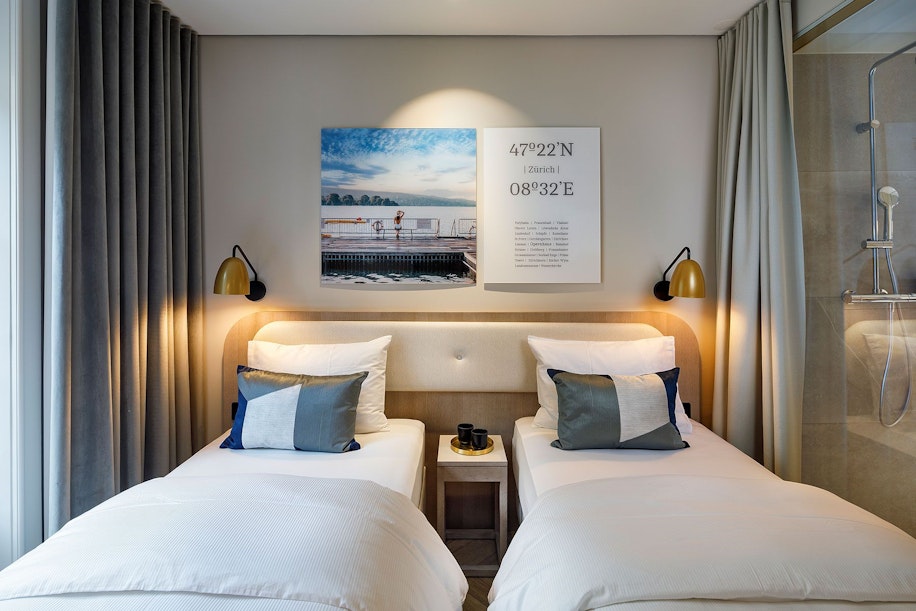 Ein modernes Hotelzimmer mit zwei sauber gemachten Einzelbetten, jeweils mit weißer Bettwäsche und Akzentkissen. Ein kleiner Tisch mit einer Tasse trennt die Betten. Darüber hängen Wandbilder und ein Koordinatendruck. Wandlampen sorgen für warmes Licht.