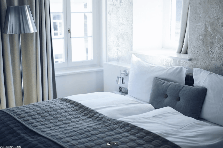Ein ordentlich gemachtes Bett mit weißen Laken, einer grauen Steppdecke und zwei Kissen in einem hellen Raum mit großen Fenstern, beigen Vorhängen, einer silbernen Stehlampe und strukturierten weißen Wänden.