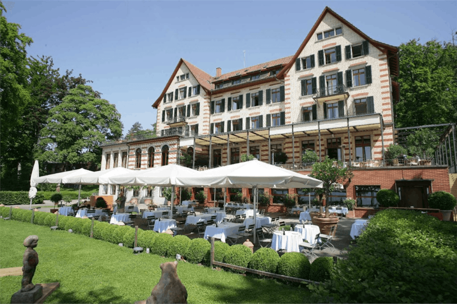 Ein großes, elegantes Hotel mit gestreifter Backsteinfassade, grünen Fensterläden und rotem Dach verfügt über einen geräumigen Essbereich im Freien, der von weißen Sonnenschirmen beschattet wird und von gepflegten Hecken und Gärten umgeben ist.