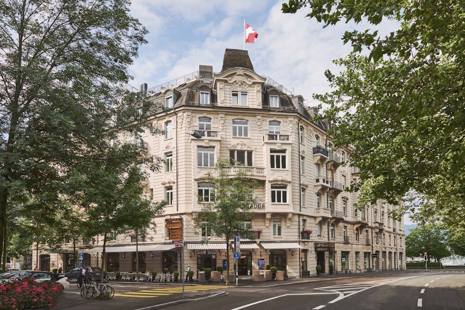 Ein historisches, reich verziertes weißes Gebäude mit einer Schweizer Flagge auf dem Dach, einer von Bäumen gesäumten Straße davor, Sitzgelegenheiten im Freien in Cafés im Erdgeschoss und ein paar in der Nähe geparkten Fahrrädern.
