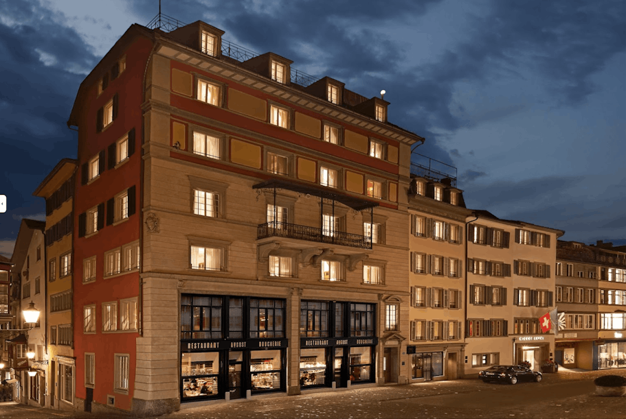 Ein hell erleuchtetes Eckgebäude mit großen Fenstern, umgeben von anderen Gebäuden im traditionellen europäischen Stil, steht in der Abenddämmerung unter einem bewölkten blauen Himmel in einer Kopfsteinpflasterstraße.