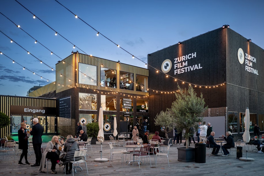 Abends sitzen Menschen an Tischen im Freien unter Lichterketten vor modernen Gebäuden mit großen Schildern des Zurich Film Festival. Einige Leute essen oder unterhalten sich, und die Atmosphäre ist lebhaft und festlich.