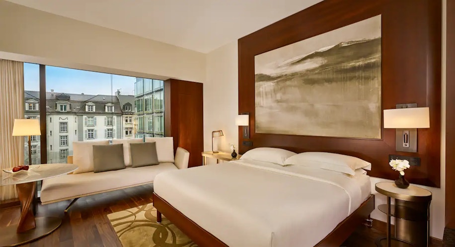 Ein modernes Hotelzimmer mit einem großen Bett, einem an der Wand hängenden abstrakten Kunstwerk, einem kleinen runden Tisch mit Stühlen, einem Sofa am Fenster und einem Blick auf die Stadtlandschaft durch raumhohe Fenster.
