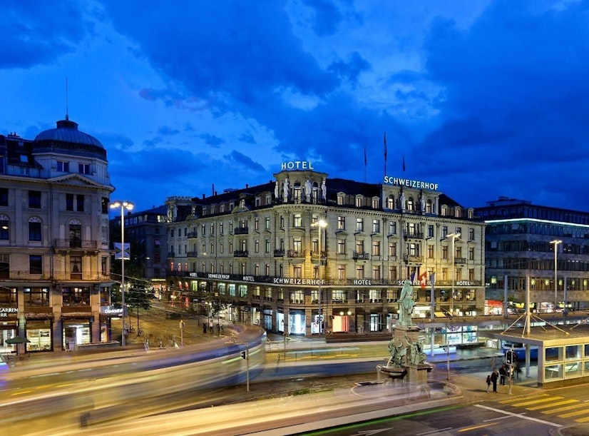 Eine Stadtszene in der Abenddämmerung mit dem beleuchteten Hotel Schweizerhof an einer belebten Straßenecke, mit verschwommenen Lichtern des vorbeifahrenden Verkehrs und einem dramatisch blauen Himmel mit Wolken darüber.