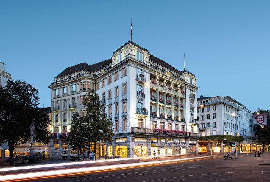 Ein großes, mehrstöckiges Hotel mit beleuchteten Balkonen und einer Schweizer Flagge auf dem Dach steht in der Abenddämmerung an einer belebten Stadtstraße, mit Lichtspuren vorbeifahrender Fahrzeuge und Geschäfte auf Straßenebene.