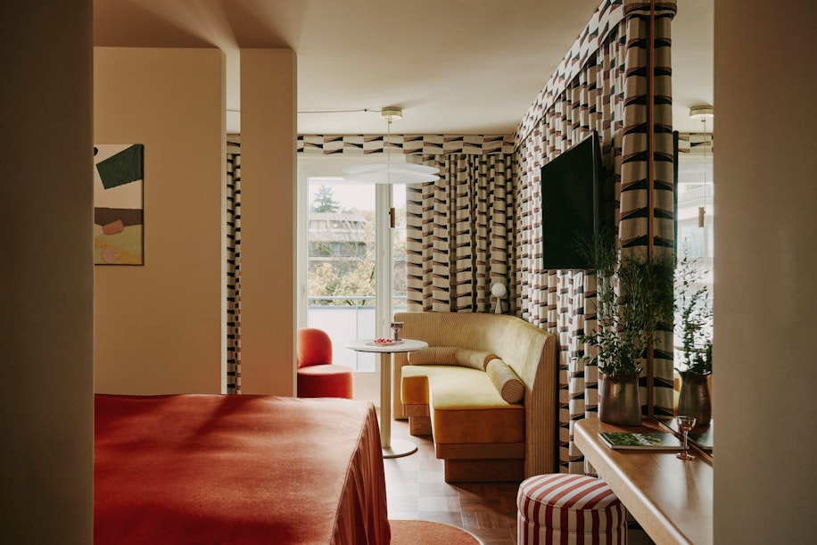 Ein modernes Hotelzimmer mit Vorhängen mit kräftigen geometrischen Mustern, einem Bett mit rot-orangefarbener Bettdecke, einem gelben Ecksofa, einem kleinen runden Tisch, gestreiften Hockern und einem Flachbildfernseher an der Wand neben einer Balkontür mit Außenlicht.
