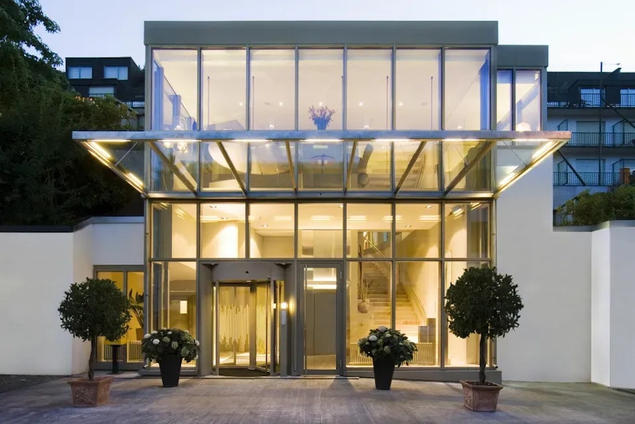 A modern glass building entrance with large windows, bright interior lighting, double glass doors, visible staircase, and potted plants on either side of the doorway.