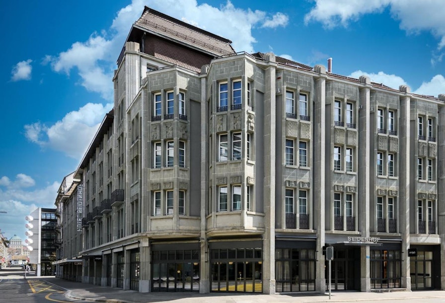 Ein großes, historisches Steingebäude mit mehreren Stockwerken und dekorativen architektonischen Details steht an einer Straßenecke unter einem blauen Himmel mit vereinzelten Wolken. Das Erdgeschoss verfügt über große Fenster und einen Eckeingang.