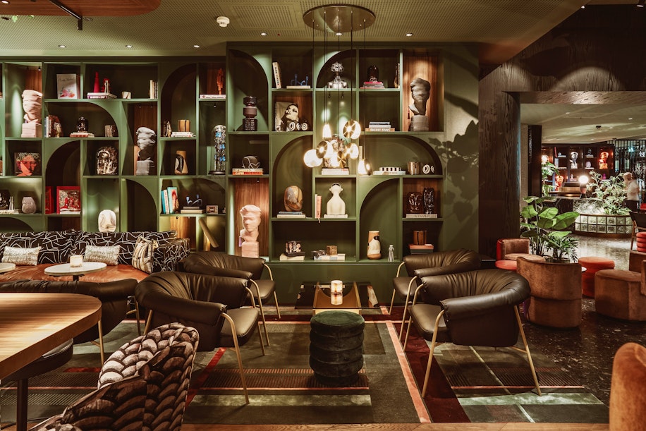 Eine stilvolle Lounge mit modernen Sesseln, runden Tischen und warmer Beleuchtung. Ein großes Wandregal präsentiert Bücher, Kunst und Skulpturen. Der Raum bietet gemütliche Sitzgelegenheiten, Dekorationsobjekte und ein schickes, einladendes Ambiente.