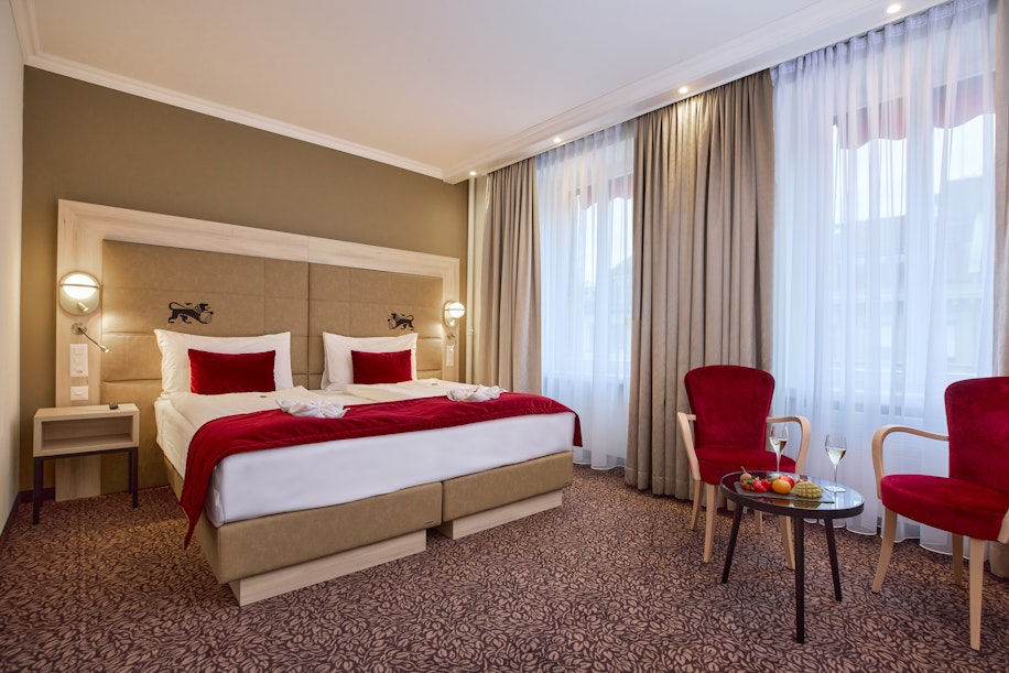 Ein Hotelzimmer mit einem großen Doppelbett, rot-weißer Bettwäsche, zwei roten Samtstühlen, einem kleinen runden Tisch mit Obst und einem Glas Wein, großen Fenstern mit transparenten Vorhängen und warmer Beleuchtung.