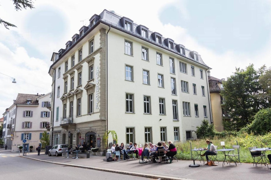 Eine Gruppe von Menschen sitzt an einem sonnigen Tag an Tischen im Freien vor einem großen, hellen, vierstöckigen Gebäude mit hohen Fenstern an einer Stadtstraße und genießt das Essen und die Unterhaltung.