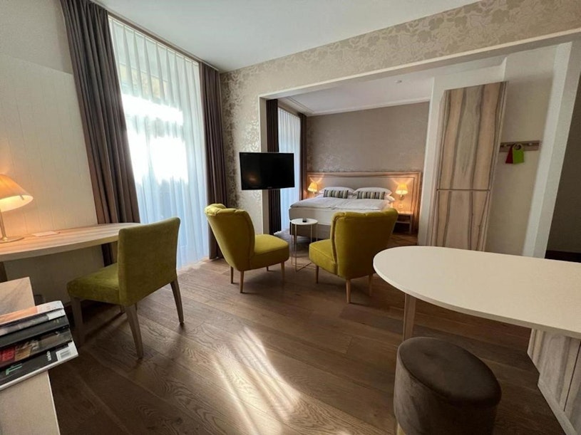 Ein modernes Hotelzimmer mit Holzboden, im Hintergrund ein Bett, zwei grüne Sessel, ein kleiner runder Tisch, ein Flachbildfernseher, ein Schreibtisch mit Lampe und große Fenster mit transparenten Vorhängen.