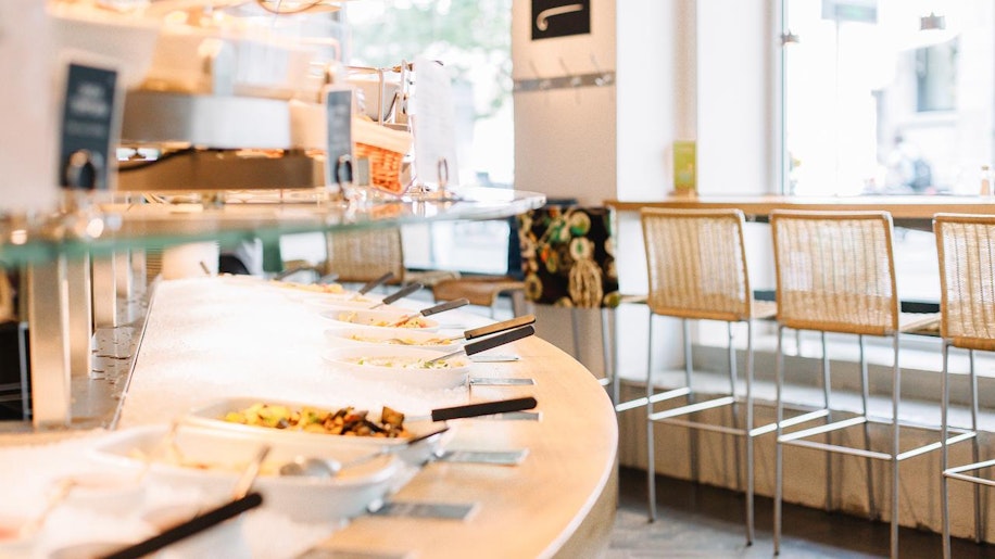 Ein helles, modernes Buffet mit mehreren Gerichten in weißen Schalen auf einer geschwungenen Theke. In jeder Schale befindet sich Servierbesteck. Im Hintergrund stehen hohe Barhocker an großen Fenstern.