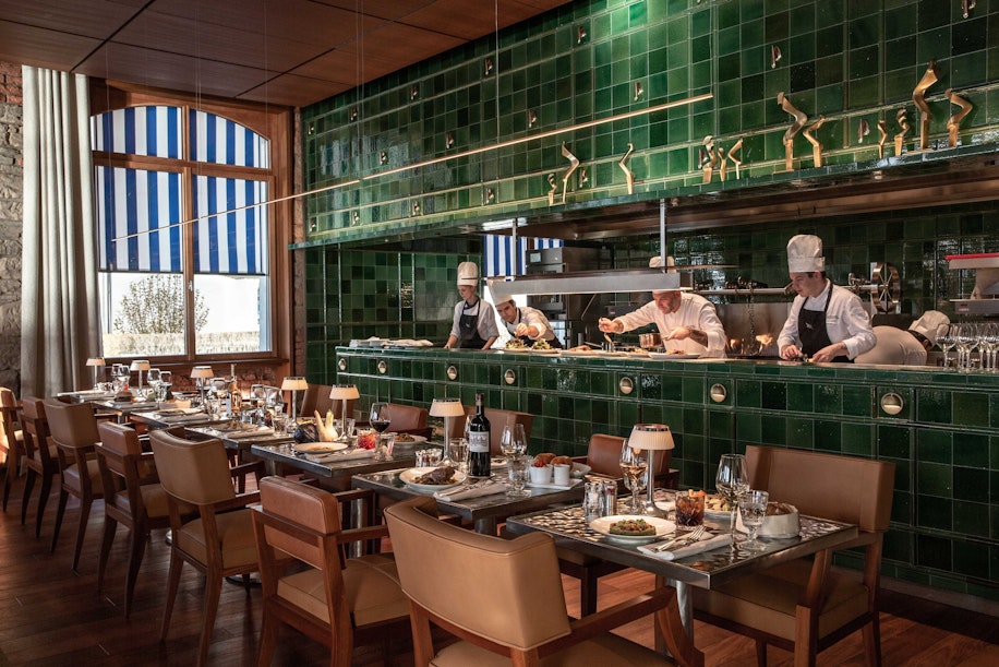 Ein stilvolles Restaurant mit grün gefliesten Wänden, Köchen in einer offenen Küche und gedeckten Tischen mit Tellern, Gläsern und Lampen. Große Fenster mit blau gestreiften Vorhängen lassen Tageslicht herein.