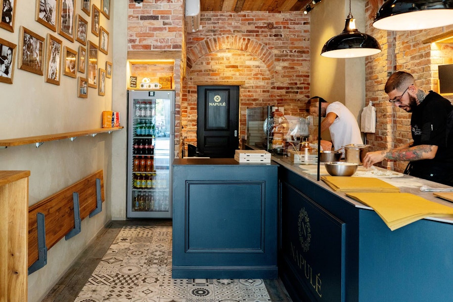 Eine gemütliche Pizzeria mit unverputzten Ziegelwänden, Vintage-Fotos, gemustertem Fliesenboden und einer blauen Theke. Zwei Mitarbeiter bereiten hinter der Theke Essen zu. Im Hintergrund steht ein Getränkekühlschrank und eine schwarze Tür.